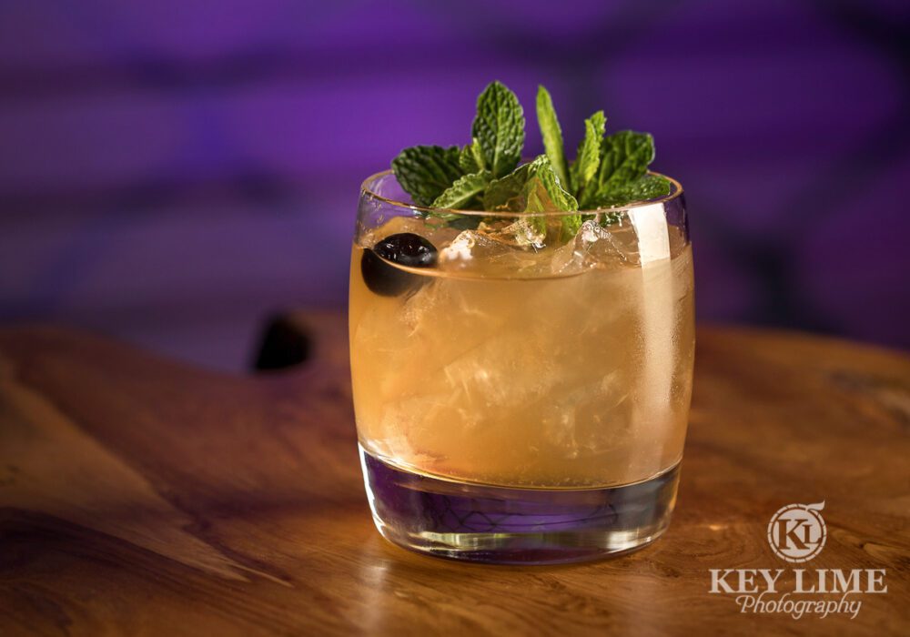 Close-up image of a cocktail on the rocks topped with basil and a black olive in front of a purple backdrop