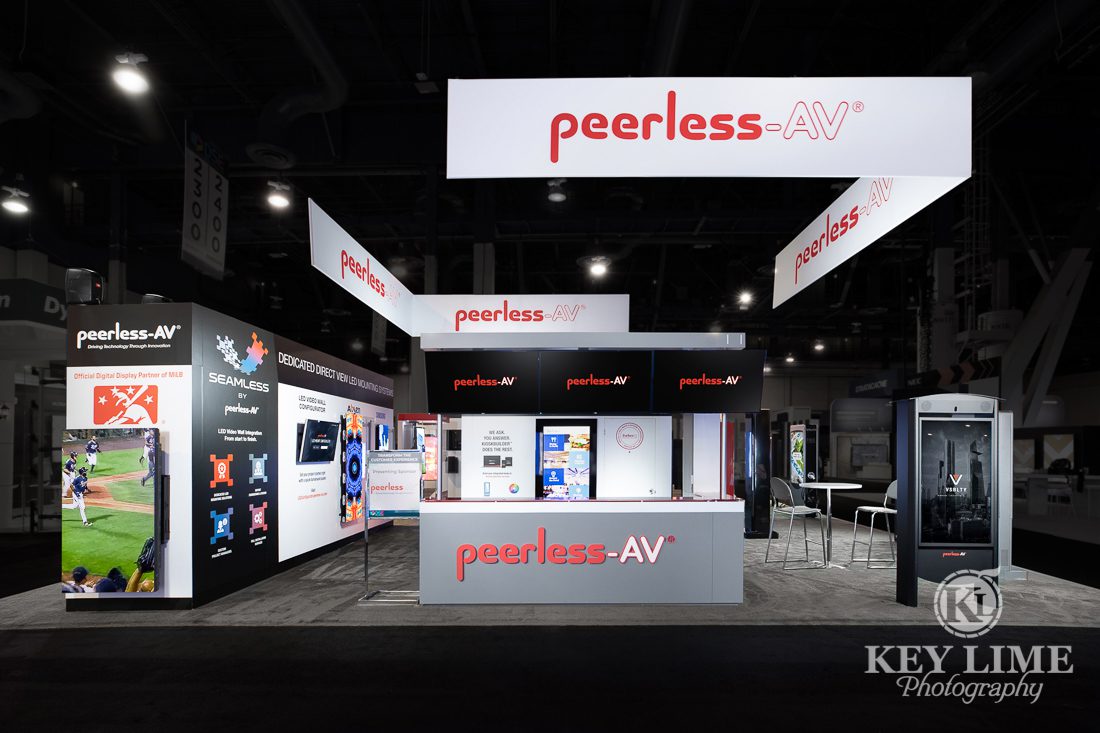 Hero trade show photographer image of white and gray booth design. Red accents, black carpet. PeerlessAV digital displays