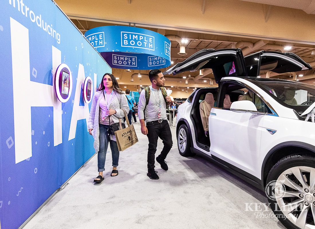 Modern trade show booth photography. Two milenials walk together near Tesla vehicle, automotive