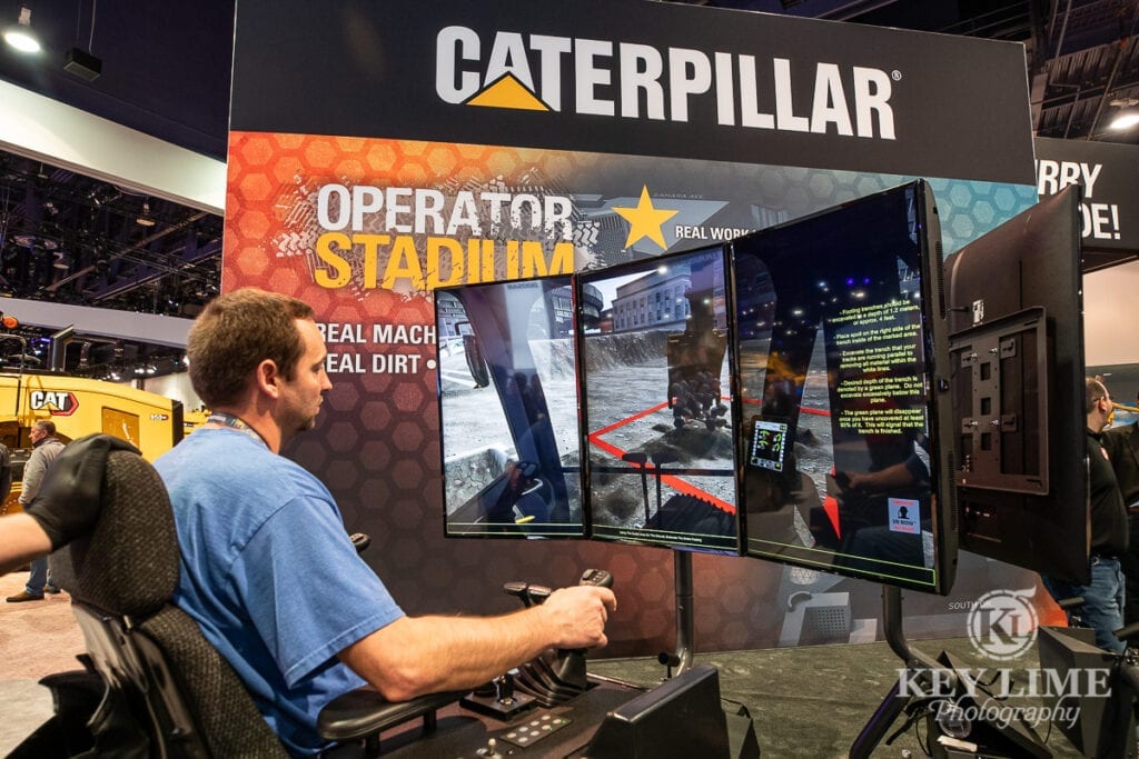 Construction photography of virtual backhoe heavy equipment video game, operator is sitting at controls that you would see in an actual vehicle and his view is simulated on a set of surrounding tv screens.