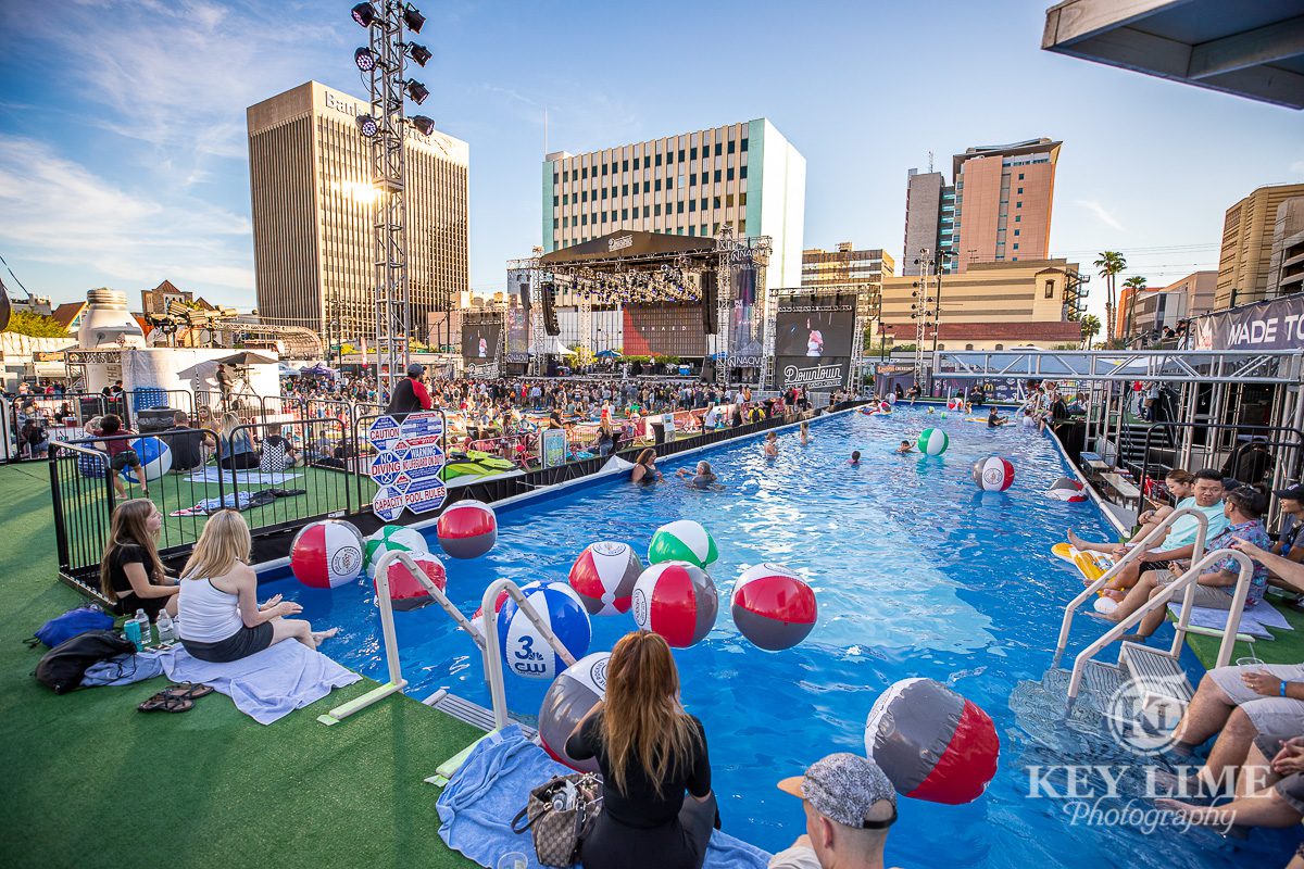 Event photography of the pool view and VIP cabanas area at Bite of Las Vegas 2019, Blue water with red and white beach balls. Girls sitting on the pool edge with the downtown Vegas city skyline in the near distance.
