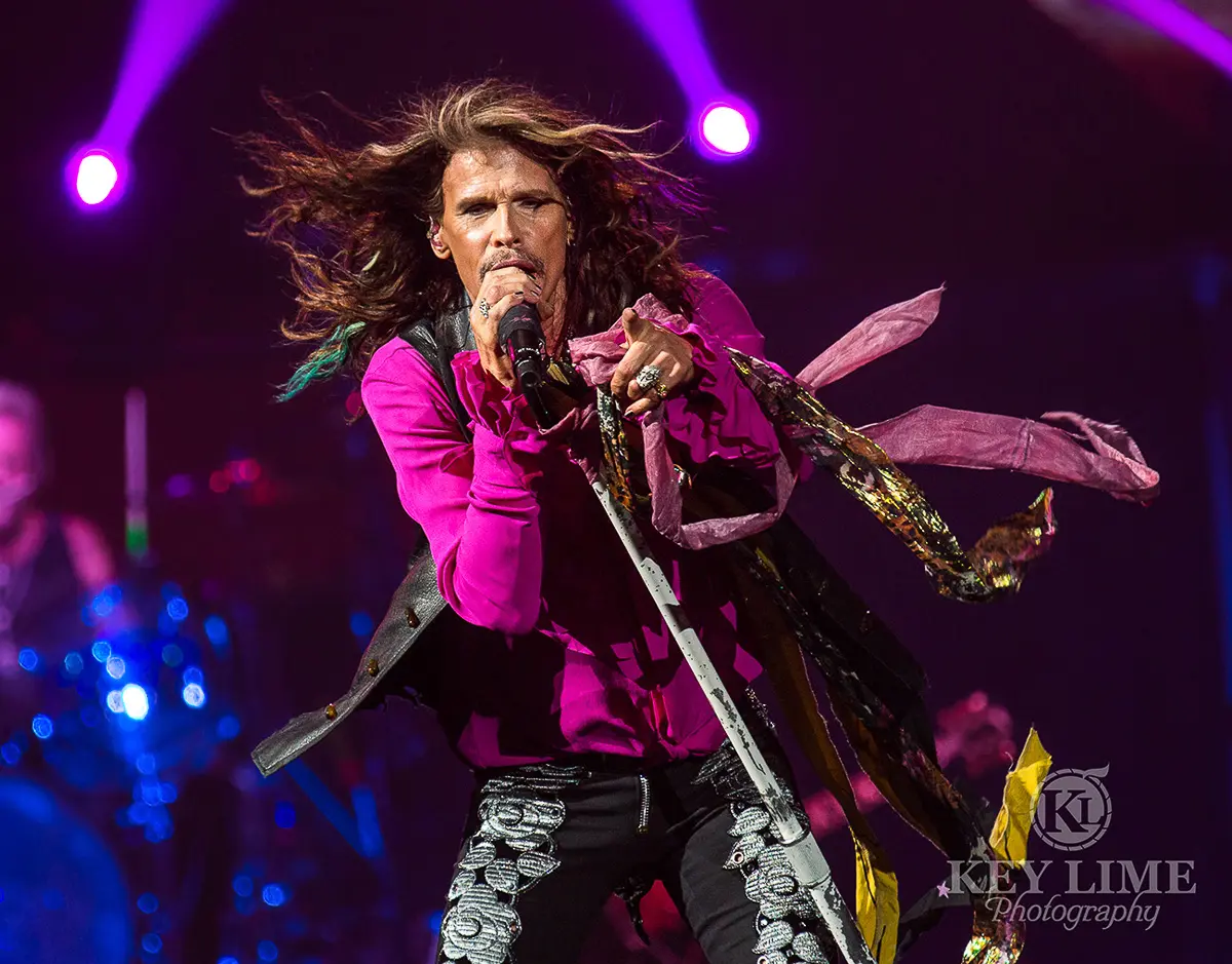 Iconic event photographer image of Steven Tyler during an Aerosmith performance. Tight, black embellished jeans with a silky magenta shirt and black vest.