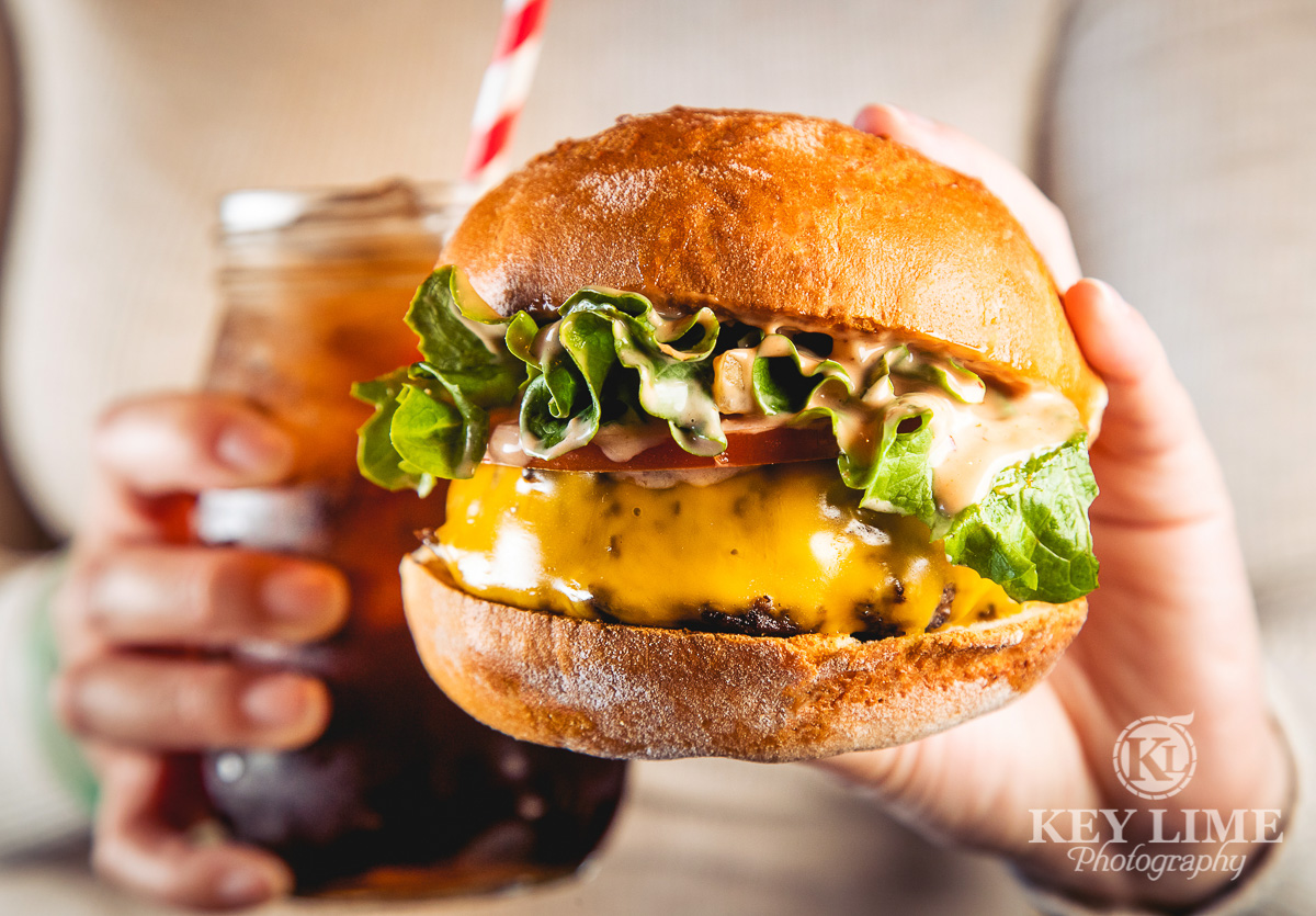 Classic cheeseburger and a Coke. Hands holding beautiful, lively burger. Ice cold glass with red and white swirly straw in the background.