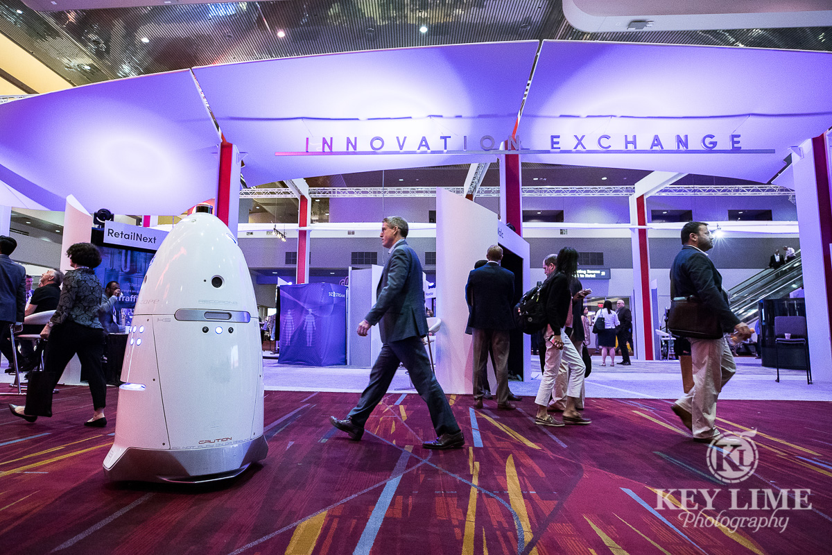 Modern trade show with automated robot greeting people. Convention call with corporate attire and white conical robot.